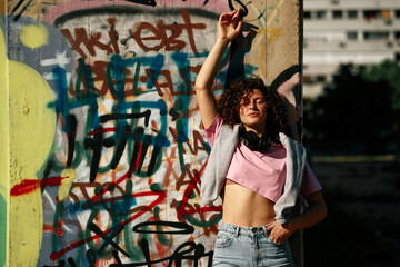 A young woman with curly hair stands against the wall and enjoying the sunny day. The woman has headphones around her neck, and on the wall, there is graffiti.