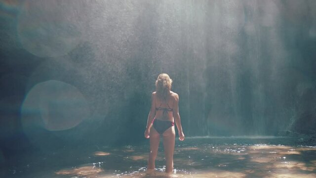 attractive woman in waterfall cave wearing bikini exploring underground cavern alone with water splashing through light rays