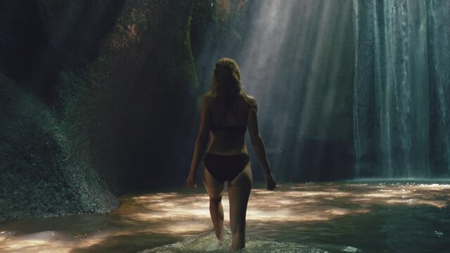 attractive woman in waterfall cave wearing bikini exploring underground cavern alone with water splashing through light rays