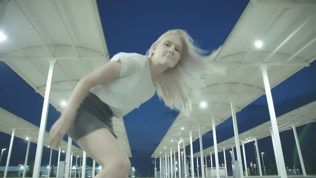 Young beautiful woman with white hair in a short skirt dances and gestures against the background of an empty car parking lot at night in bright light