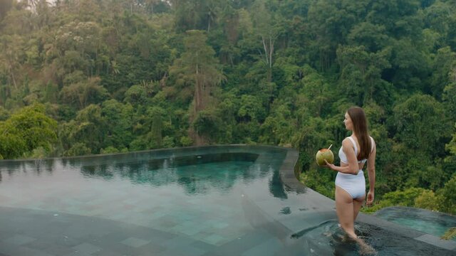 beautiful woman by infinity pool drinking coconut cocktail enjoying luxury lifestyle on exotic summer vacation at hotel spa with view of tropical jungle