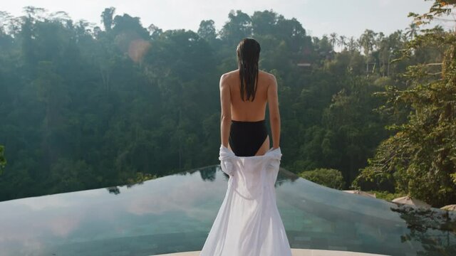 sexy woman walking into infinity pool undressing to swim enjoying luxury lifestyle on exotic summer vacation at hotel spa with view of tropical jungle 4k