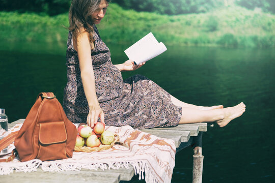Pregnant woman sitting on wooden pier near lake reading book and eating apples. Outdoor picnic near water. Wellness and time to relax alone.