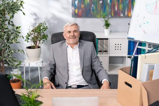 Sympathetic Handsome Older Man Boss Manager Of Company Is Sitting Dressed In Suit On A Comfortable Chair At Desk, Businessman Finished Work At Computer Relaxes Looks Into Camera With Smile