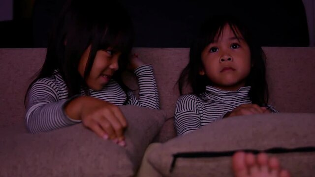 Cute little children sitting on sofa and watching cartoons on TV in the living room late in evening. Happy Asian sisters watch TV at home and spend time together on vacation.