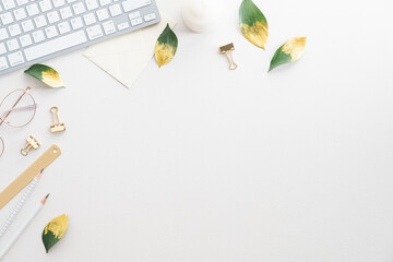 Feminine desk table with keyboard, green leaves, office supplies on white background. Flat lay, top view, copy space.