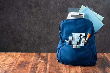 Blue backpack with stationery over wooden table and black background.