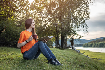 A young beautiful woman in bright clothes is sitting on the green grass and enjoying reading a book in nature. Concept of a pleasant pastime, rest, relaxation