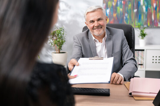 Older Boss In Suit Sits In Office Chair At Desk, Conducts Job Interview, Man Is Pleased With Woman, Hands Her Employment Contract To Sign, Smiles Both Hold Up Pad Of Document