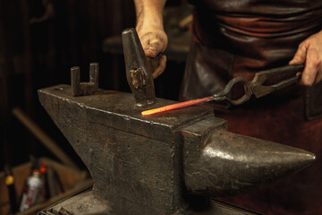 Close-up working powerful hands of male blacksmith forge an iron product in a blacksmith. Hammer, red hot metal and anvil. Concept of labor, retro professions