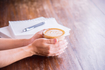 Hands holding a cup of coffee with foam and notepaper on wooden table