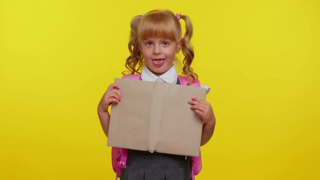 Smiling school girl hiding behind a book, laugh fooling around showing tongue making playful silly facial expressions, showing tongue, grimacing over yellow studio background. Back to school concept