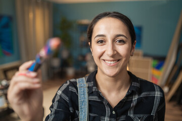 Portrait of a smiling, happy lady stretching out her hand in which she holds a brush dirty with oil paint, watercolor, in the background living room in the evening, canvases, easels