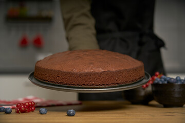 A proud cook woman shows the homemade chocolate cake she has just baked in her kitchen. She will decorate the sweet bakery with blackberries, red currants, powdered sugar and dark chocolate.