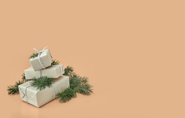 Small Christmas gift boxes wrapped in craft paper with green fir tree decorated branches on solid beige background. Modern minimal xmas presents pastel backdrop with copy space. New Year celebration 