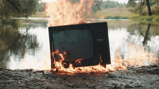Retro vintage TV flares up and burns with fire next to the river. Concept of post apocalypse, surrealism and the fire hazard of old worn out equipment