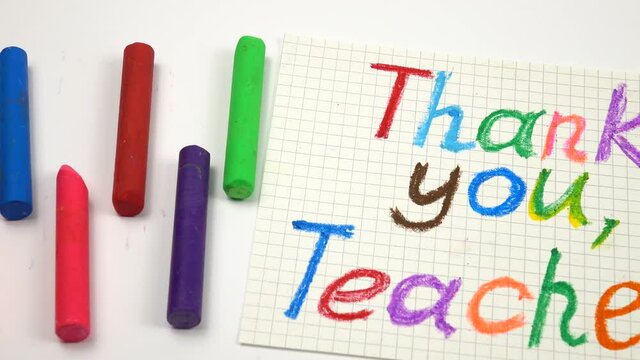 A greeting handwritten card with the gratitude to the teacher in honor of teacher's day, pieces of the colorful chalks lie on a white table