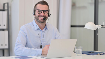 Middle Aged Man with Headset Smiling at Camera while using Laptop 