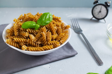 Raw fusilli in a white bowl on a gray napkin with a clock on a gray background. Time to eat pasta. Selective focus. Horizontal orientation. Whole grain pasta.