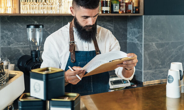 Handsome Barista Doing an Inventory of the Products in a Cafe. 
Serious waiter with a beard standing in the bar counter and using clipboard for counting stock in the restaurant.