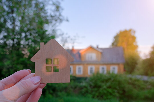 female hand holds a small toy wooden house. In the background is a real residential building. horizontal photo. Idea - a dream of owning a home, buying, selling, renting real estate.