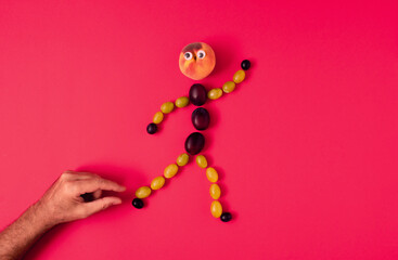 A human figure made of grape grains, plums and peach runs away from the male hand. Minimalistic creative funny autumn concept, flat lay composition on pastel red background