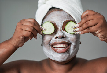 Young african woman with skin care cream holding fresh cucumber slices in front of her eyes -...