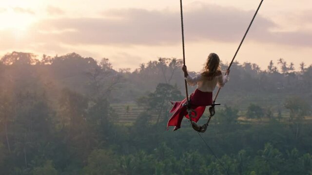 travel woman swinging over tropical rainforest at sunrise female tourist sitting on swing with scenic view enjoying freedom on vacation having fun holiday lifestyle slow motion
