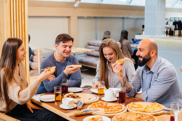 A group of young cheerful friends is sitting in a cafe talking and eating pizza. Lunch at the pizzeria.