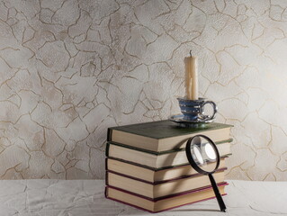 a candle made of wax stands on a stack of old thick books above a magnifying glass