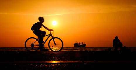 Obraz premium Silhouette of woman riding bicycle along the coastal road during the sunset.