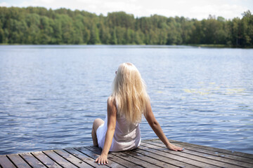 Young beautiful blonde hair woman sitting on the wooden pier and looking at the lake