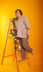 a charming woman of American appearance of 50 years in a beige suit stands on the stairs in flowers on a yellow background. concept of elderly joyful
