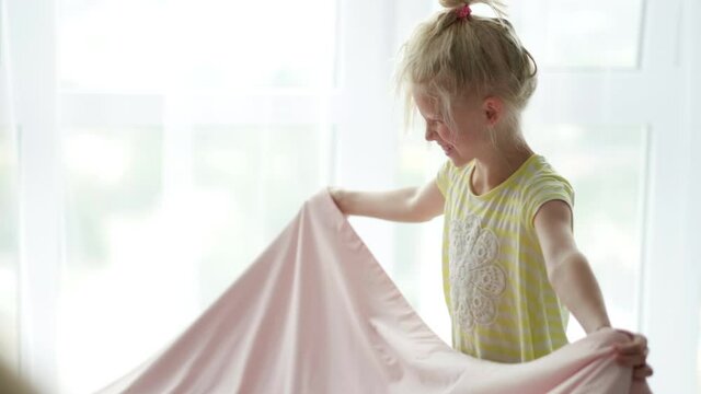 Smiling little blonde girl making bed in morning at home. Child waves the blanket to spread it. Fresh bedclothes. Everyday routines.