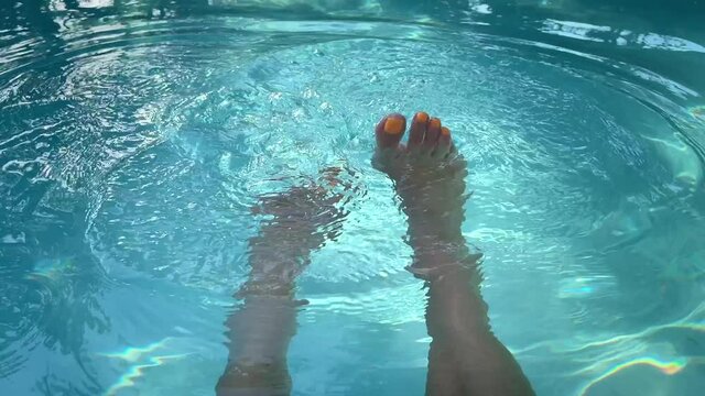 Female legs with orange nail paint splashing in a swimming pool