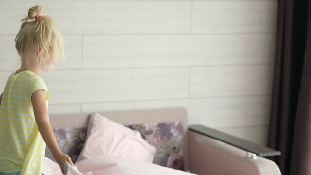 Smiling little blonde girl making bed in morning at home. Child waves the blanket to spread it. Fresh bedclothes. Everyday routines.