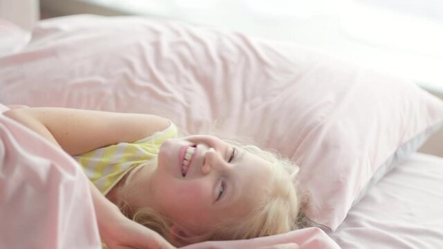 Portrait handsome cute little blonde girl having fun in bed in the morning. Adorable kid spinning, laughing, lying upside down.