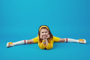 Cute girl child doing splits, stretching exercise, straight angle pose, working out wearing yellow outfit, indoor full length, blue studio background - obrazy, fototapety, plakaty