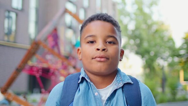 Portrait of joyful cute schoolboy standing outdoors and fixing his backpack on the shoulders. Confident African american student near school smiling to camera.