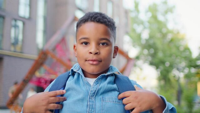 Portrait of joyful cute schoolboy standing outdoors and putting on backpack. Male African american student near school smiling to camera.