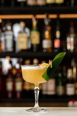 Glass with yellow lemon cocktail decorated with dried pineapple and mint on the blurred bar counter background.