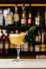 Glass with yellow lemon cocktail decorated with dried pineapple and mint on the blurred bar counter background.