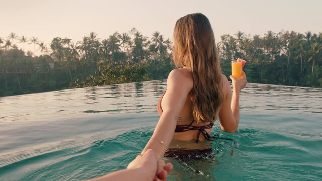 travel couple holding hands woman leading boyfriend in swimming pool enjoying luxury honeymoon on exotic vacation at hotel spa with view of tropical jungle at sunrise