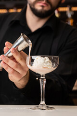 male bartender pouring to the measuring glass cup with ice cream an alcoholic drink from steel jigger on the bar counter on the blurred background
