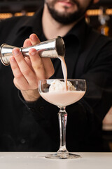 male bartender pouring to the measuring glass cup with ice cream an alcoholic drink from steel jigger on the bar counter on the blurred background