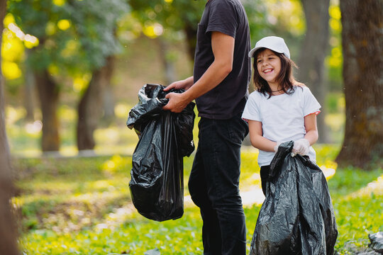 Photo Of A Smiling Beautiful Girl And Adult Man Volunteer Gathering Rubbish Together Outdoors Using Big Black Garbage Bags.