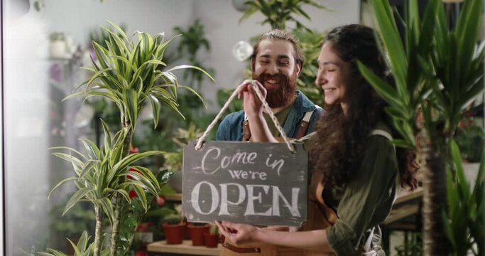 Flower shop employees hanging welcoming wooden open sign on glass entrance door. Happy employees starting new workday 4k footage