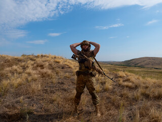 Fototapeta premium Photo of a mercenary sniper in camouflage clothes under the scorching sun. He stands with a rifle and surveys the area.