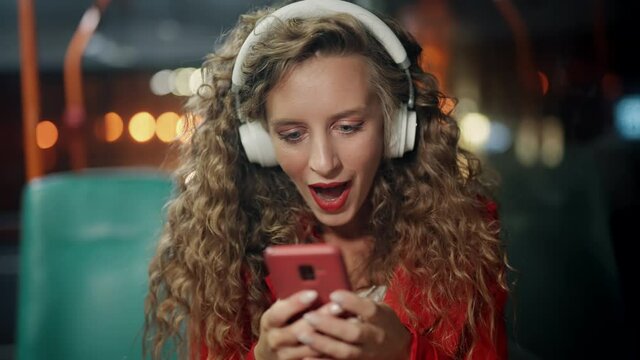 A curly woman is joying good message or news on smartphone in public transport. A girl got a positive answer or approval. Surprising and getting shocked. Concept of victory and gain. Night time.
