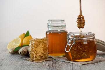 Honey in a jars, piece of bee comb and honey dipper on light background. Next to it is sliced lemons and ginger. Home treatment for flu. Honey harvesting concept.